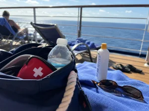 A small, white first aid kit with a red cross icon on its lid, resting on a wooden deck next to a pair of sunglasses and a white beach hat, with a blue ocean and distant land visible in the background