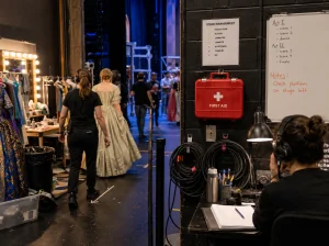 Un botiquín de primeros auxilios de teatro abierto que revela suministros médicos. La caja blanca tiene una cruz roja y la palabra "First Aid" impresa en su tapa. Vendas, gasas, esparadrapo y toallitas antisépticas son visibles en el interior.